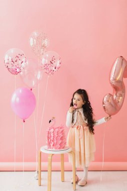 Pembe elbiseli sevimli küçük kız flamingo doğum günü pastasının yanında poz veriyor ve pembe arka planda beşinci doğum gününü kutluyor.