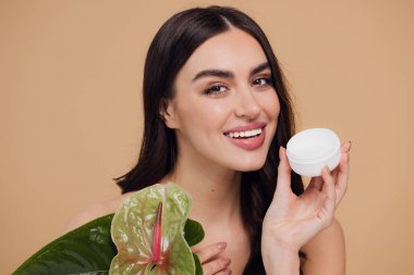 Yeşil çiçeğin yanındaki çekici bir kadının portresi sorun için yüz kremi sürüyor. Bej arka planda merhem var. Refah konsepti