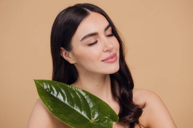 Temiz, parlak yüzlü, bej arka planda yeşil yapraklı genç bir kadın. Spa, yüz bakımı, güzellik kozmetolojisi. Doğal kozmetik konsepti.