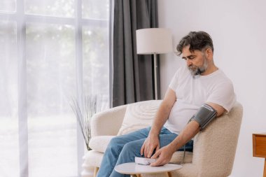 Evdeki kanepede oturmuş, tansiyonunu ölçen, gri saçlı, son sınıf öğrencisi. Modern tonometre kullanan emekli, hipertansiyon deneyimi yaşayan, kopyalama alanı