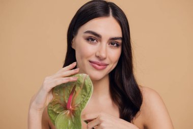 Bej renkli stüdyo arka planında izole edilmiş çiçekle poz veren çekici genç bir kadının güzel portresi.