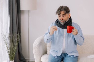 Yüksek ateşi olan yaşlı bir adam, termometresini dikkatlice kontrol ediyor ve evdeyken bir fincan çay içiyor. Etrafı çeşitli ilaçlarla çevrili bir battaniyeye sarılmış ve kendini pek iyi hissetmiyor gibi görünüyor..