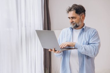 Oturma odasında dizüstü bilgisayar kullanarak gülümseyen olgun bir adam. Mutlu kendinden emin yetişkin erkek evde dinleniyor bilgisayar üzerinde çalışıyor, klavyede daktilo yazıyor, boşluğu kopyalıyor