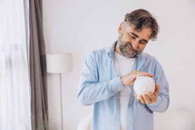 Sakallı yaşlı adam beyaz bir kumbara gösteriyor. Tasarruf temaları, emeklilik ve fotokopi alanı ile finansal planlama öneriyor.