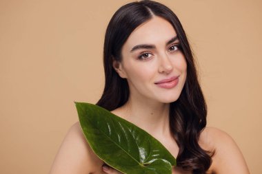 Temiz, parlak yüzlü, bej arka planda yeşil yapraklı genç bir kadın. Spa, yüz bakımı, güzellik kozmetolojisi. Doğal kozmetik konsepti.