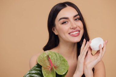 Yeşil çiçeğin yanındaki çekici bir kadının portresi sorun için yüz kremi sürüyor. Bej arka planda merhem var. Refah konsepti