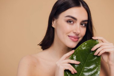 Temiz, parlak yüzlü, bej arka planda yeşil yapraklı genç bir kadın. Spa, yüz bakımı, güzellik kozmetolojisi. Doğal kozmetik konsepti.
