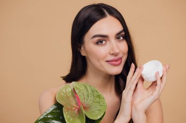 Yeşil çiçeğin yanındaki çekici bir kadının portresi sorun için yüz kremi sürüyor. Bej arka planda merhem var. Refah konsepti