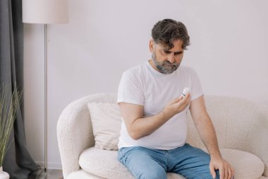 Oturma odasındaki koltukta oturan yaşlı adam kan basıncını cihazla ölçüyor. Yaşlı erkek hapları tutuyor ve uyuşturucu bırakıyor. Sağlık ve öz bakım kavramı, kopyalama alanı