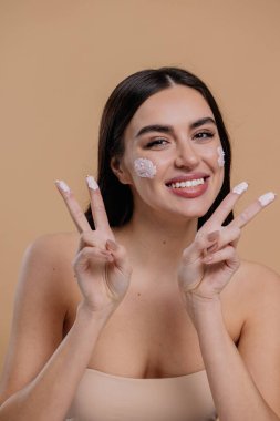Kadın güzel, mutlu yüz, krem sürerek ve cildini temizlerken parmakları, tırnağı ve saçı, güzel saçı, vücudu ve bej arka planda güzellik konsepti.