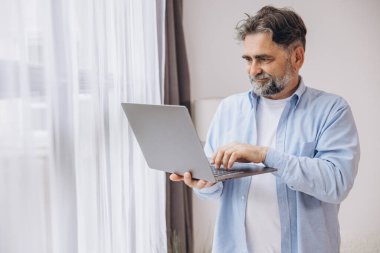 Oturma odasında dizüstü bilgisayar kullanarak gülümseyen olgun bir adam. Mutlu kendinden emin yetişkin erkek evde dinleniyor bilgisayar üzerinde çalışıyor, klavyede daktilo yazıyor, boşluğu kopyalıyor