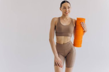 Bej rengi spor giysili zayıf bir kadının portresi spor salonu için turuncu köpük silindirli güvenli bir şekilde duruyor, fotokopi alanı ile beyaz üzerinde poz veriyor, sağlık ve refahı destekliyor..