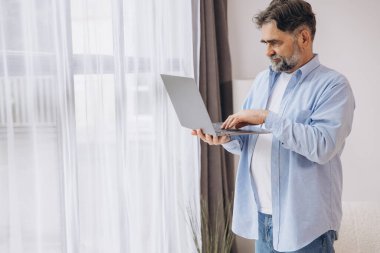 Oturma odasında dizüstü bilgisayar kullanarak gülümseyen olgun bir adam. Mutlu kendinden emin yetişkin erkek evde dinleniyor bilgisayar üzerinde çalışıyor, klavyede daktilo yazıyor, boşluğu kopyalıyor