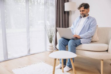 Oturma odasında dizüstü bilgisayar kullanarak gülümseyen olgun bir adam. Mutlu kendinden emin yetişkin erkek evde dinleniyor bilgisayar üzerinde çalışıyor, klavyede daktilo yazıyor, vücut uzunluğu, kopyalama alanı