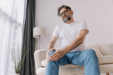 Ev, diz ağrısı ve koltukta eklem iltihabı, kemik erimesi ve fibromiyalji olan yaşlı bir adam. Rahatlama, kas gerilimi ve kaza ya da emeklilikte yaralanma için eklem masajı yapan yaşlı erkek.