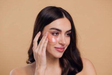 Göz bantları. Güzellik konsepti. Güzel bir model gözlerine pembe hidro jel sürerek bej arka planda gülümsüyor.