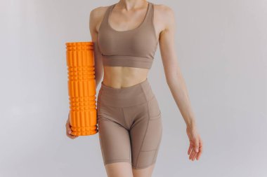 Sıska bir kadının bej rengi spor giysili portresi spor salonu için turuncu köpük silindirli güvenli bir şekilde duruyor. Fotokopi alanı ile beyaza poz veriyor, sağlık ve sağlığı destekliyor..