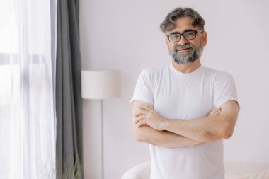 Mutlu, kendinden emin, gözlüklü, kolları bağlı yaşlı bir adam. Yaşlı olgun gülümseyen 60 yaşındaki yakışıklı adam modern evdeki oturma odasında poz veriyor. Portre.