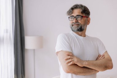 Mutlu, kendinden emin, gözlüklü, kolları bağlı yaşlı bir adam. Yaşlı olgun gülümseyen 60 yaşındaki yakışıklı adam modern evdeki oturma odasında poz veriyor. Portre.