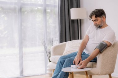 Evdeki kanepede oturmuş, tansiyonunu ölçen, gri saçlı, son sınıf öğrencisi. Modern tonometre kullanan emekli, hipertansiyon deneyimi yaşayan, kopyalama alanı