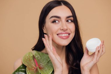 Yeşil çiçeğin yanındaki çekici bir kadının portresi sorun için yüz kremi sürüyor. Bej arka planda merhem var. Refah konsepti