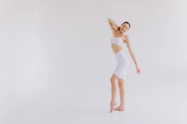 Beyaz arka planda beyaz spor giysili zayıf bir kadın var. Düz göbekli, kaslı, zayıf, çekici bir kadın. Uzayı metin için kopyala. Vücut bakımı, sağlıklı ve sportif yaşam, saç aldırma, yoga konsepti