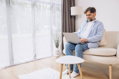 Oturma odasında dizüstü bilgisayar kullanarak gülümseyen olgun bir adam. Mutlu kendinden emin yetişkin erkek evde dinleniyor bilgisayar üzerinde çalışıyor, klavyede daktilo yazıyor, vücut uzunluğu, kopyalama alanı