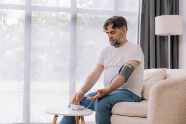 Evdeki kanepede oturmuş, tansiyonunu ölçen, gri saçlı, son sınıf öğrencisi. Modern tonometre kullanan emekli, hipertansiyon deneyimi yaşayan, kopyalama alanı