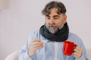 Yüksek ateşi olan yaşlı bir adam, termometresini dikkatlice kontrol ediyor ve evdeyken bir fincan çay içiyor. Etrafı çeşitli ilaçlarla çevrili bir battaniyeye sarılmış ve kendini pek iyi hissetmiyor gibi görünüyor..