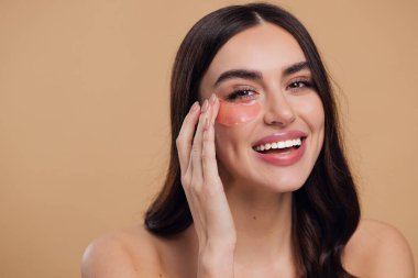 Göz bantları. Güzellik konsepti. Güzel bir model gözlerine pembe hidro jel sürerek bej arka planda gülümsüyor.