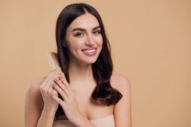 Çıplak omuzlarını bayanla su kuyusu-damat rüya gibi esmer saç o ahşap fırça pastel bej renkli arka plan üzerinde izole ile saç tarama büyüleyici portre yapmak beyaz ışınlama dişleri kazma dişli bir gülümseme