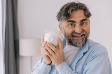 Sakallı yaşlı adam beyaz bir kumbara gösteriyor. Tasarruf temaları, emeklilik ve fotokopi alanı ile finansal planlama öneriyor.