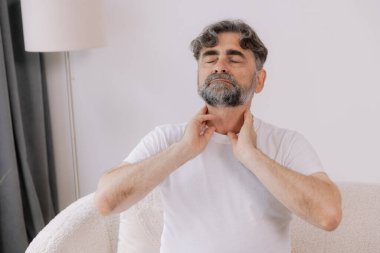 Gri saçlı adamın boğazı ağrıyormuş. Yaşlı bir erkek koltukta oturup elini boğaz iltihabından tutmuş ve boğazı ağrıyormuş..