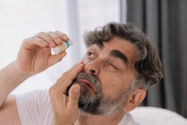 Close-up photo of an older gray-faced man who drips eye drops with medicine for pain and fatigue, sitting on the couch at home. clipart