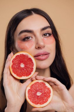 Güzel, saf, nazik bir kadının çıplak, soyutlanmış bir kamera gibi duran görüntüsü. Göz altı pembe bantlarla cildine dikkat et ve bej arka planda yarım greyfurtlarla fotokopi çektir.