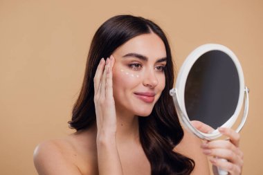 Göz Cildi Bakıcı Kadın. Göz altı derisine göz kremi süren ve bej arka planda aynaya bakan güzel genç bir kadın. Yaş Karşıtı Tedavi Konsepti. Yüksek Kalite