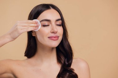 Güzel esmer, yarı çıplak bir kadın pamukla yüzünden makyaj malzemelerini çıkarıyor. Mutlu milenyum bayanı pamuk bezle deriyi temizliyor, bej rengi stüdyo arka planı, vesikalık resmi, kopyalama alanı