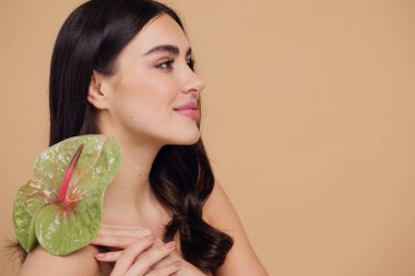 Bej renkli stüdyo arka planında izole edilmiş çiçekle poz veren çekici genç bir kadının güzel portresi.