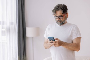 Beyaz tişörtlü, elinde cep telefonu olan ve kameraya gülümseyen yaşlı gri saçlı bir adamın portresi..