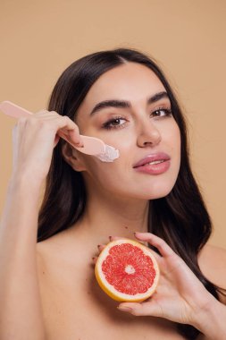 Yarı çıplak kadın, güzellik için yüz ve fırça kremi, kuaför ve cilt bakımı bej arka planda. Mutlu model gülümsüyor ve elinde greyfurt, yüz losyonu ve dermatoloji için makyaj malzemesi tutuyor.