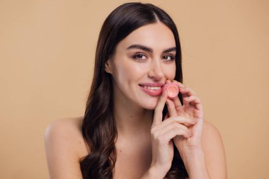 Gülümseyen bir kadının güzel portresi dudaklarını bej arka planda önemser. Makyajlı kozmetik ürünleri uygulayan esmer bir kadın. Dudak nemlendirici ya da parlatıcı kullanan manken hidrasyon, beslenme, yumuşatma merhemi