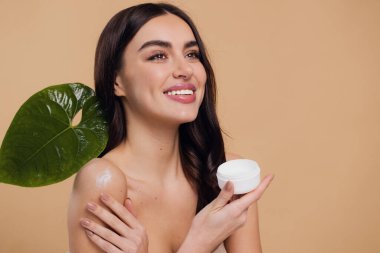 Gülen, yarı çıplak, gülümseyen ve bej arka planda izole edilmiş yüz kremi süren güzel bir kadının güzel portresini kapat.