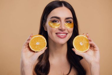 Güzel, saf, kibar bir kadının çıplak, izole bir şekilde duran görüntüsü. Gözlerinin altında altın bantlarla cildine dikkat et. Bej arka planda yarım portakal ile fotokopi alanı tut.