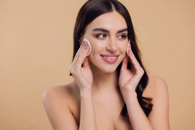 Güzel esmer, yarı çıplak bir kadın pamukla yüzünden makyaj malzemelerini çıkarıyor. Mutlu milenyum bayanı pamuk bezle deriyi temizliyor, bej rengi stüdyo arka planı, vesikalık resmi, kopyalama alanı