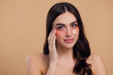 Göz bantları. Güzellik konsepti. Güzel bir model gözlerine pembe hidro jel sürerek bej arka planda gülümsüyor.