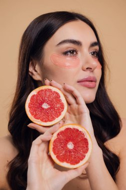 Güzel, saf, nazik bir kadının çıplak, soyutlanmış bir kamera gibi duran görüntüsü. Göz altı pembe bantlarla cildine dikkat et ve bej arka planda yarım greyfurtlarla fotokopi çektir.