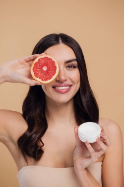 Kadın, doğal cilt bakımı için nemlendirici krem ve greyfurt ve bej renkli stüdyo arka planına karşı C vitamini. Sağlıklı organik ürünler için meyve ve krem ya da losyon tutan mutlu kadın portresi
