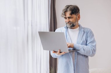 Oturma odasında dizüstü bilgisayar kullanarak gülümseyen olgun bir adam. Mutlu kendinden emin yetişkin erkek evde dinleniyor bilgisayar üzerinde çalışıyor, klavyede daktilo yazıyor, boşluğu kopyalıyor
