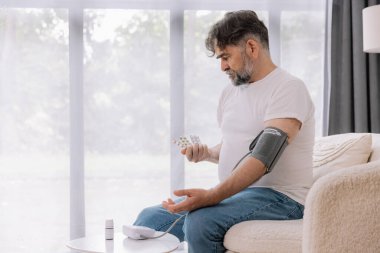 Evdeki kanepede oturmuş, tansiyonunu ölçen, gri saçlı, son sınıf öğrencisi. Modern tonometre kullanan emekli, hipertansiyon deneyimi yaşayan, kopyalama alanı