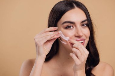 Göz bantları. Güzellik konsepti. Güzel bir model gözlerine pembe hidro jel sürerek bej arka planda gülümsüyor.